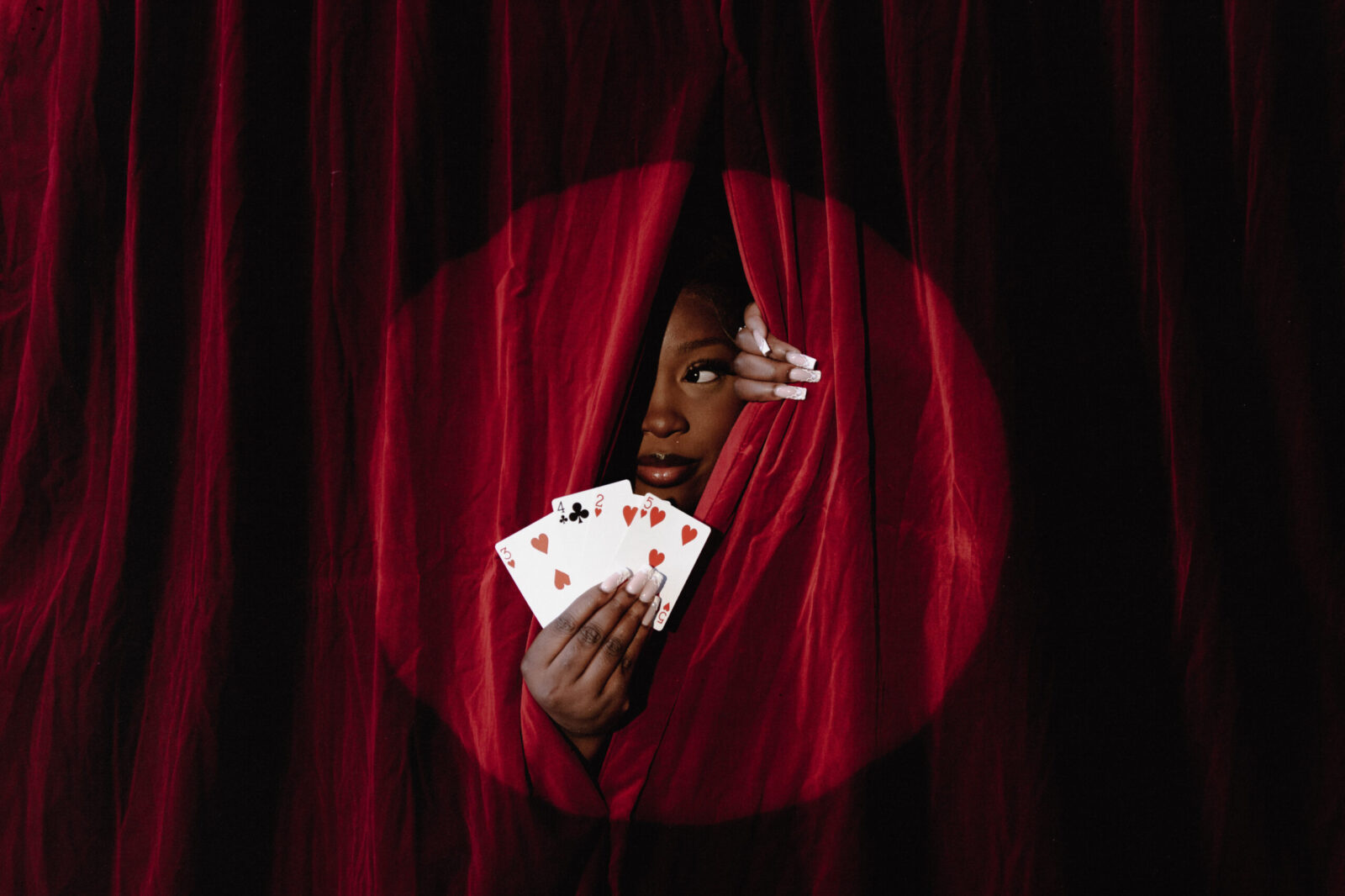 Devaura peeking out from red velvet curtains holding 3 playing cards