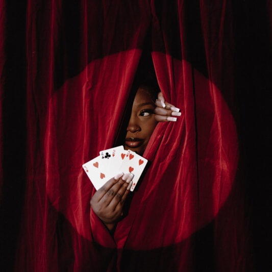 Devaura peeking out from red velvet curtains holding 3 playing cards